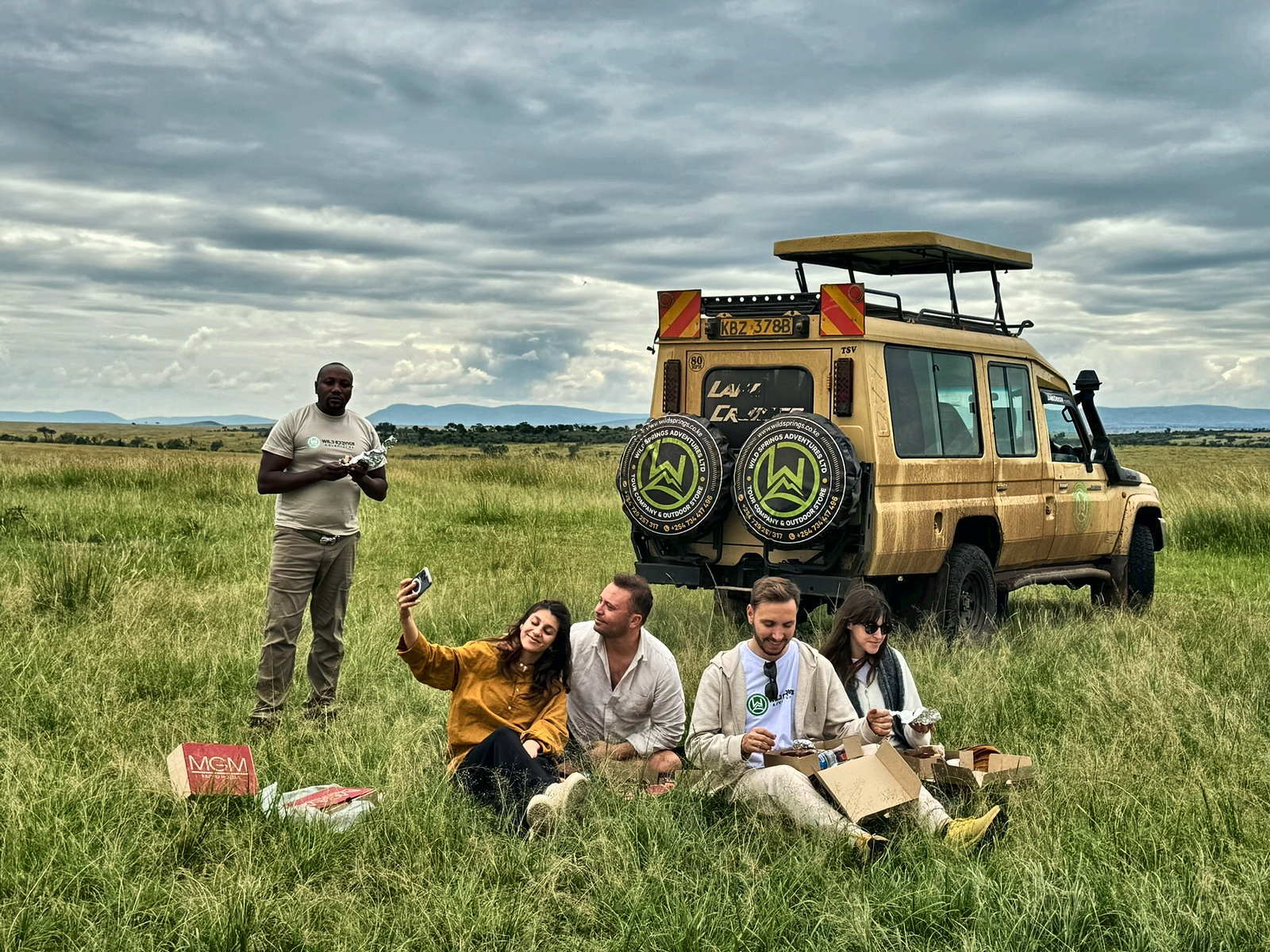 Wild Springs Adventures guests share a bush picnic beside their private 4x4 Land Cruiser on the open grassland of the Maasai Mara, Kenya, with their guide